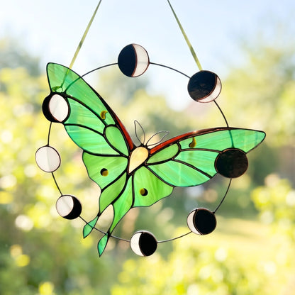A stained glass suncatcher shaped like a green butterfly with a circular moon phase design, hanging outdoors against a blurred green garden background.