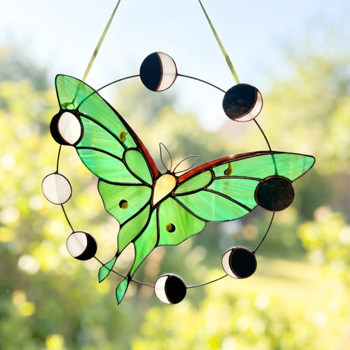 A stained glass suncatcher shaped like a green butterfly with a circular moon phase design, hanging outdoors against a blurred green garden background.