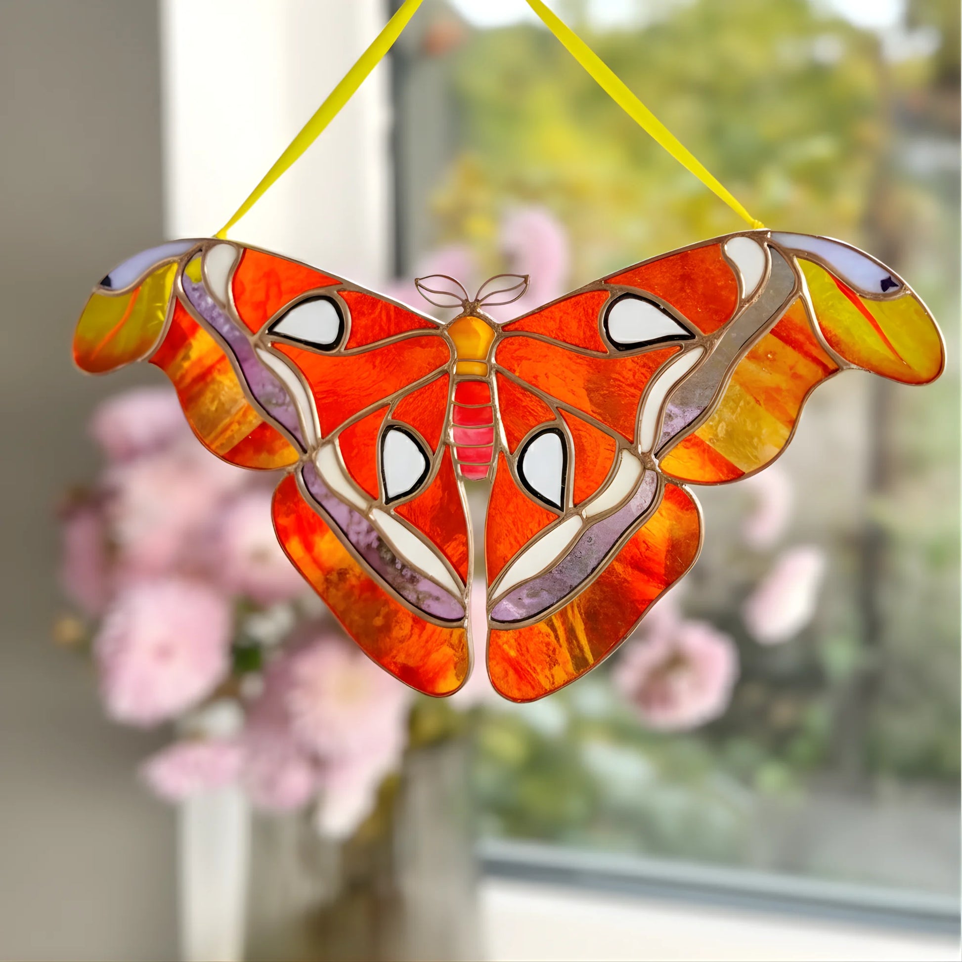 A stained glass moth suncatcher with vibrant orange, red, yellow, and white patterns, suspended by a yellow string in front of a softly blurred window with pink flowers.