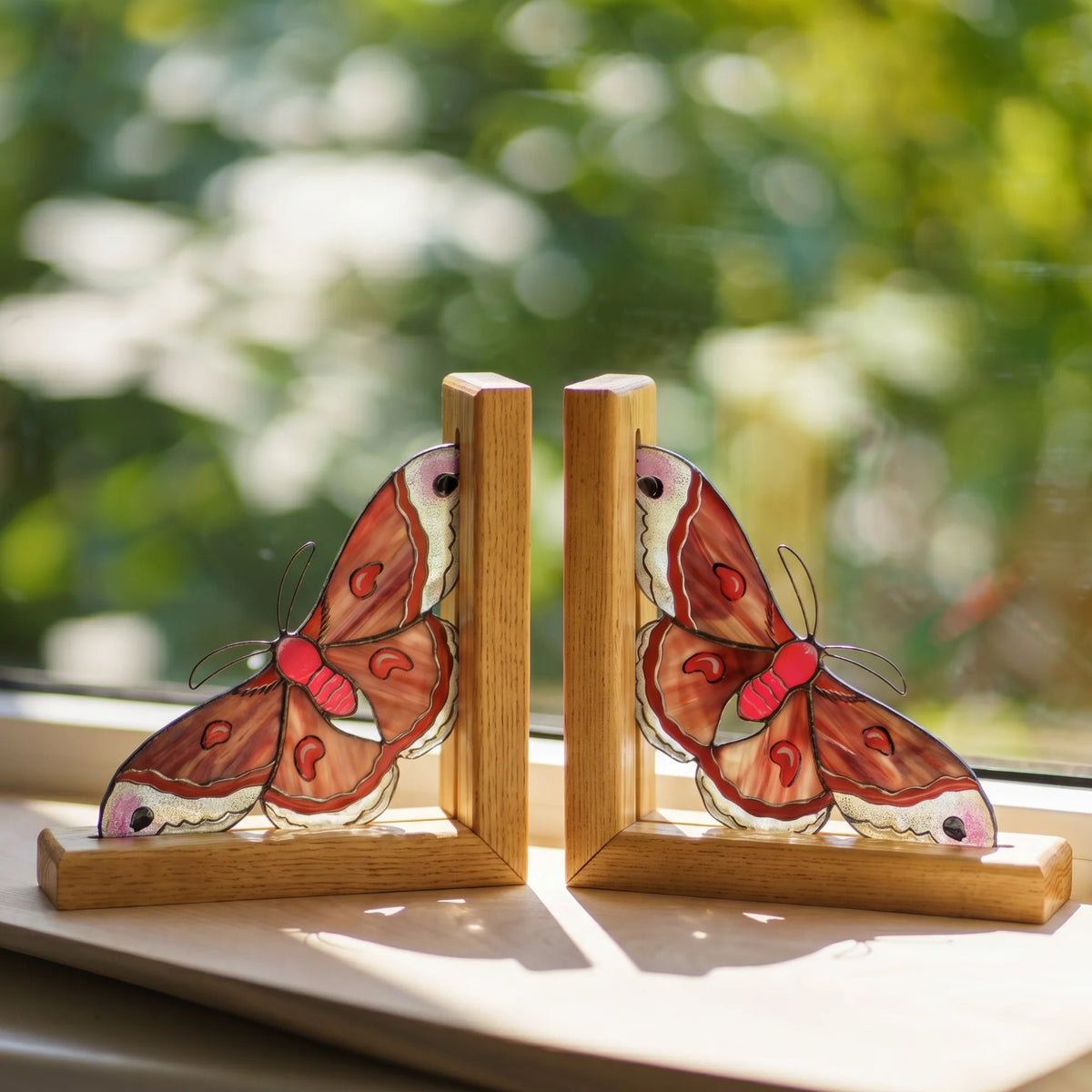 Two wooden cecropia moth bookends with detailed stained-glass style butterfly wings in brown, red, and cream, displayed on a windowsill with a blurred green garden background.