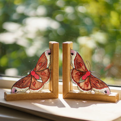 Two wooden cecropia moth bookends with detailed stained-glass style butterfly wings in brown, red, and cream, displayed on a windowsill with a blurred green garden background.
