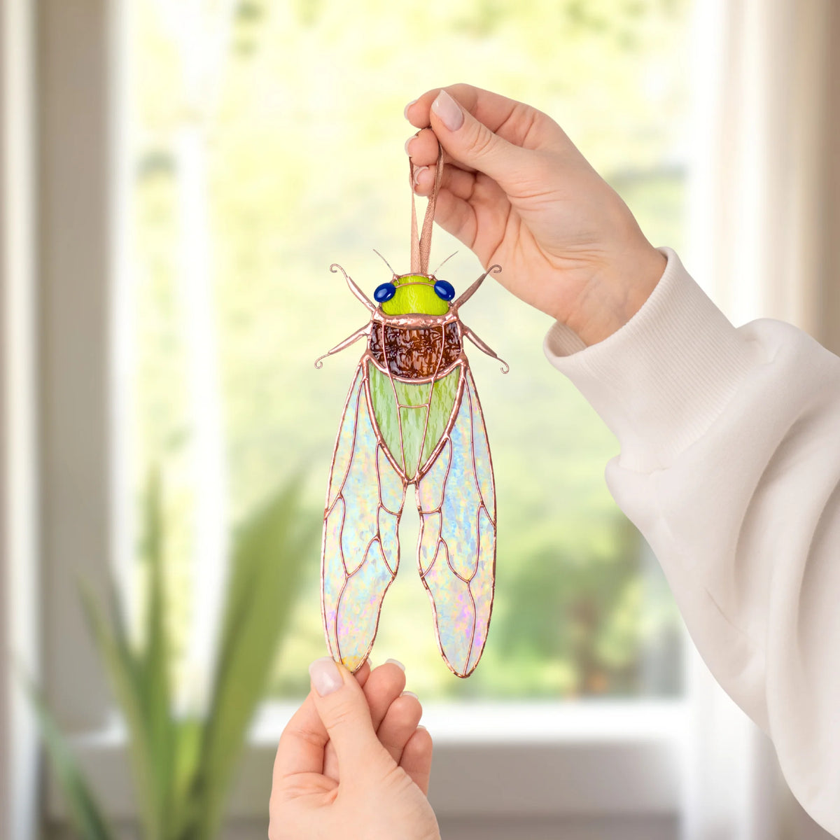 Two hands holding a green stained glass cicada window hanging up to a bright window, the iridescent stained glass wings glowing with copper veins