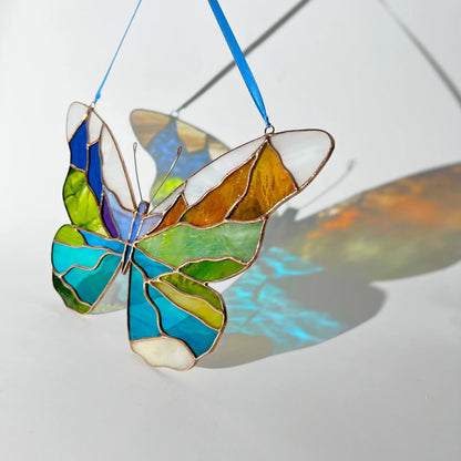 A stained glass butterfly ornament with a blue hanging ribbon, featuring segments of blue, green, yellow, orange, and white, displayed on a light surface with a colorful shadow.