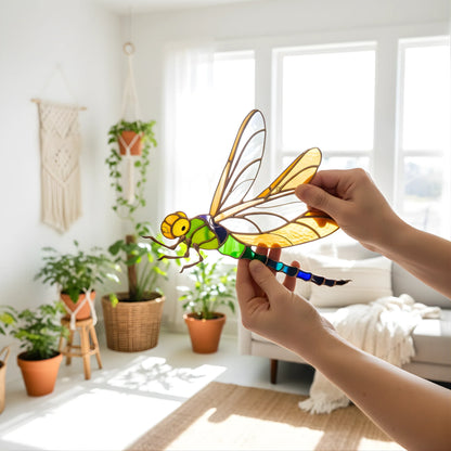 A person's hands holding a stained glass dragonfly suncatcher in a brightly lit, modern living room with potted plants and a sofa in the background.