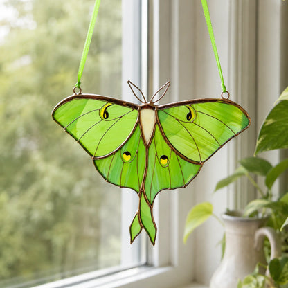 A stained glass luna moth suncatcher hanging in front of a window, with green ribbons at the top and yellow and black eyes on the wings.