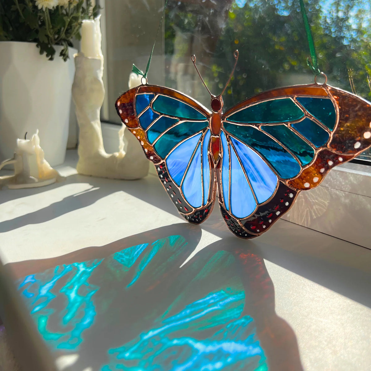 A stained glass morpho butterfly suncatcher with iridescent blue and teal wings and copper-gold outlines, hanging on a windowsill with sunlight casting colorful reflections on the windowsurface.