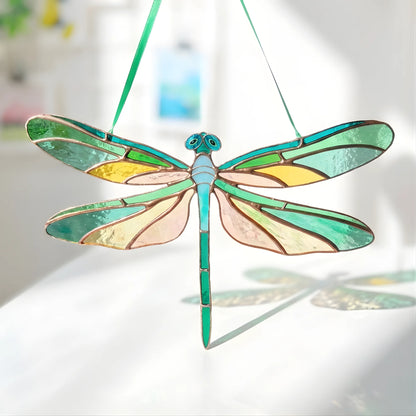 A stained-glass dragonfly suncatcher with a green body and wings featuring yellow, green, and pale pink sections, suspended by a green string against a bright, softly blurred background.