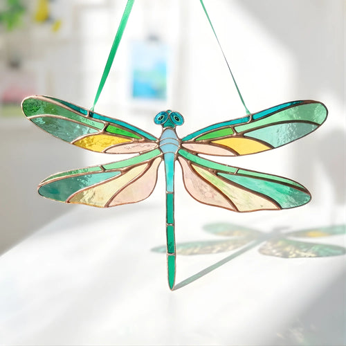 A stained-glass dragonfly suncatcher with a green body and wings featuring yellow, green, and pale pink sections, suspended by a green string against a bright, softly blurred background.