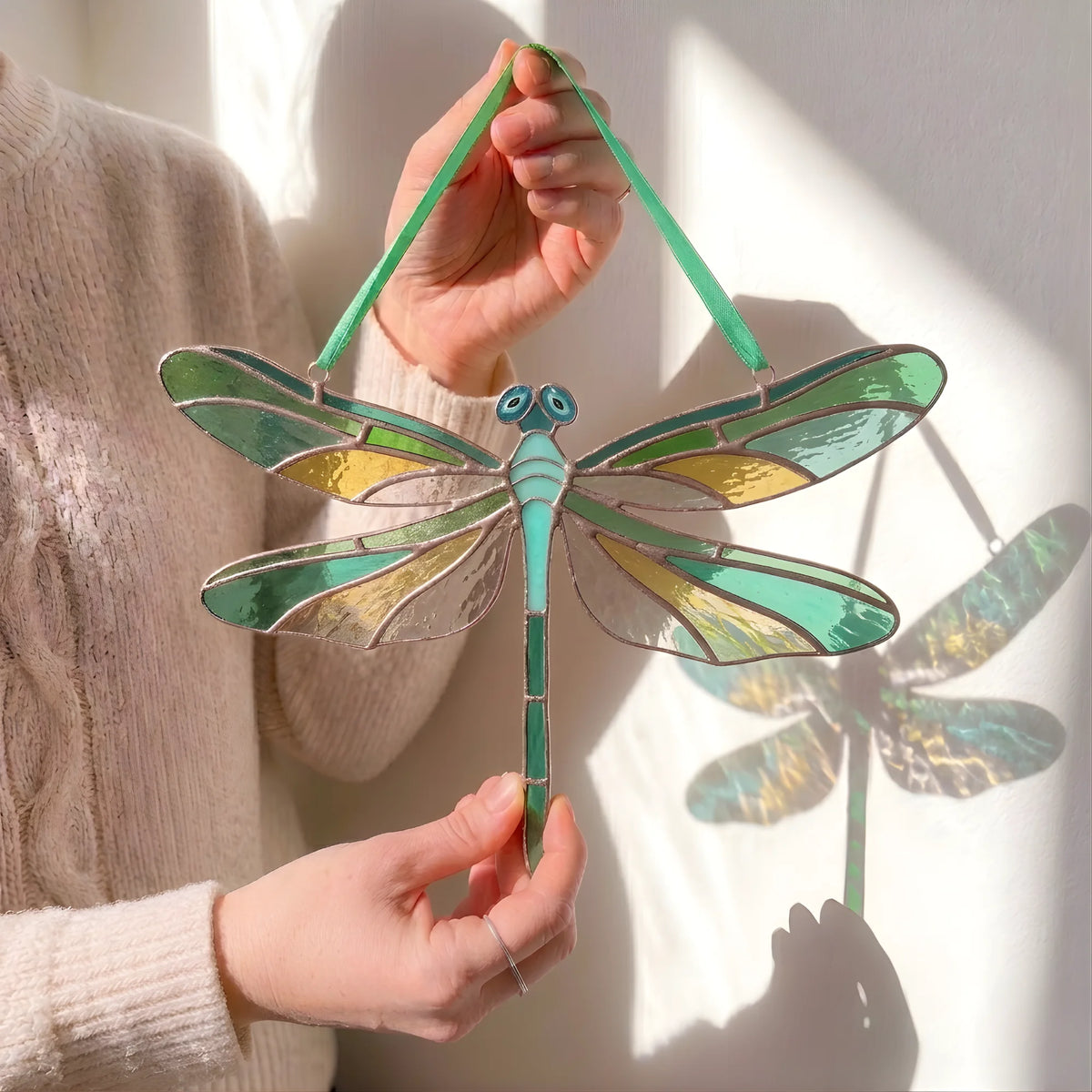 A person holds a stained-glass dragonfly suncatcher with green, teal, and gold sections outlined in dark metal, against a sunlit white wall with a dragonfly shadow cast behind it.