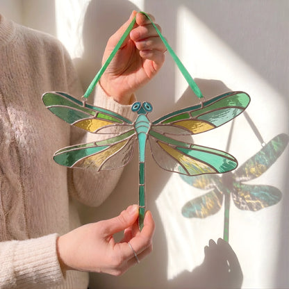 A person holds a stained-glass dragonfly suncatcher with green, teal, and gold sections outlined in dark metal, against a sunlit white wall with a dragonfly shadow cast behind it.