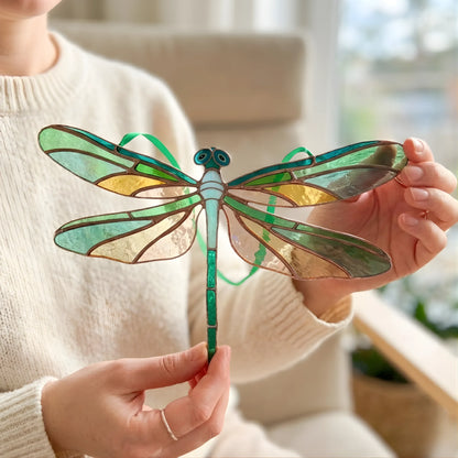 A person holds a stained glass dragonfly ornament with green, teal, and gold-toned glass panels and a green hanging ribbon, shown in a softly lit indoor setting.