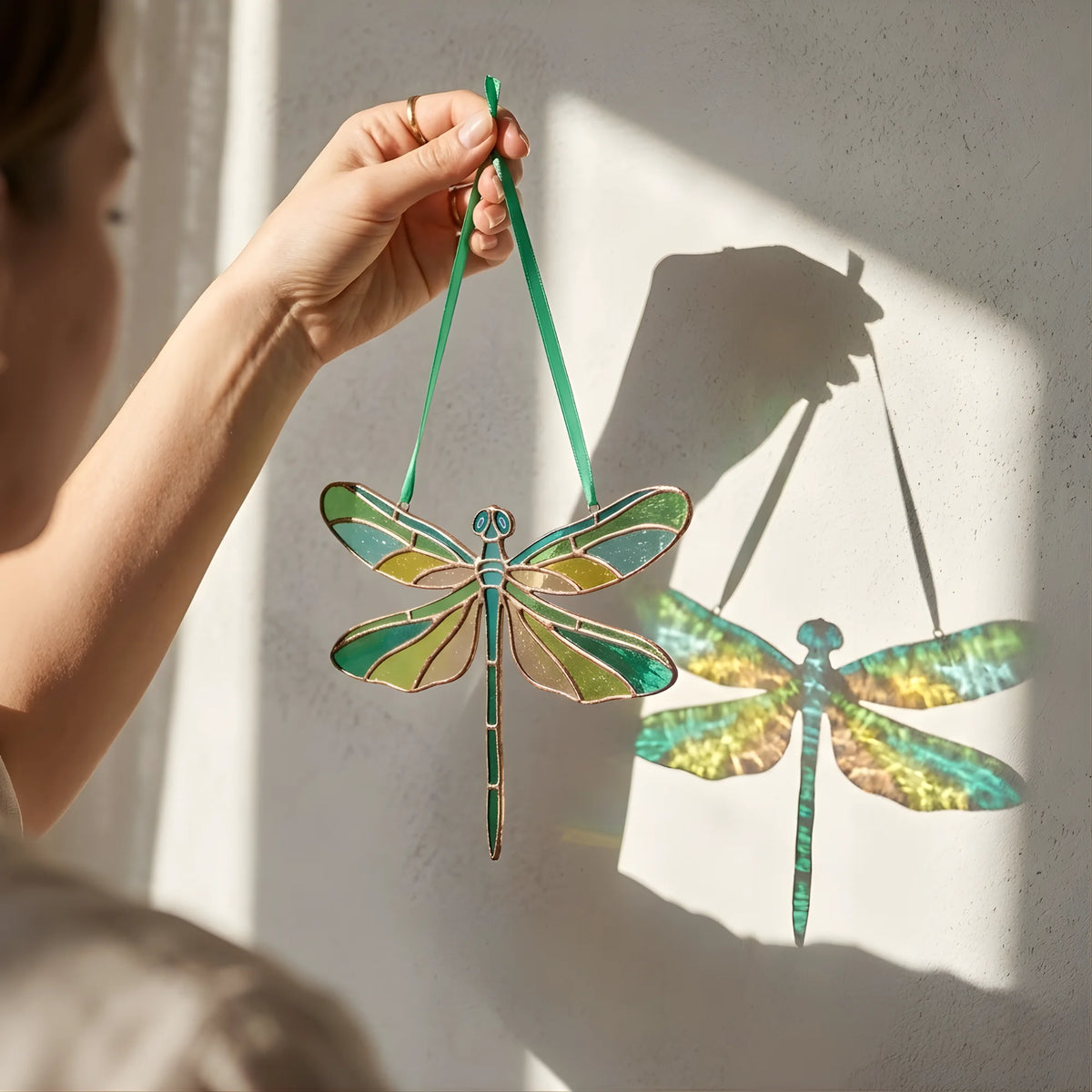 A person holds a stained-glass dragonfly ornament with a green hanging ribbon against a sunlit wall, casting a shadow of the dragonfly on the wall.