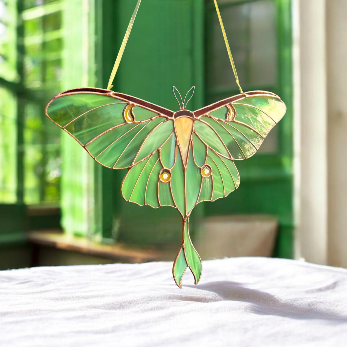 A stained glass suncatcher shaped like a luna moth with iridescent green wings and gold accents, hanging on a white fabric-covered surface with a green window in the background.