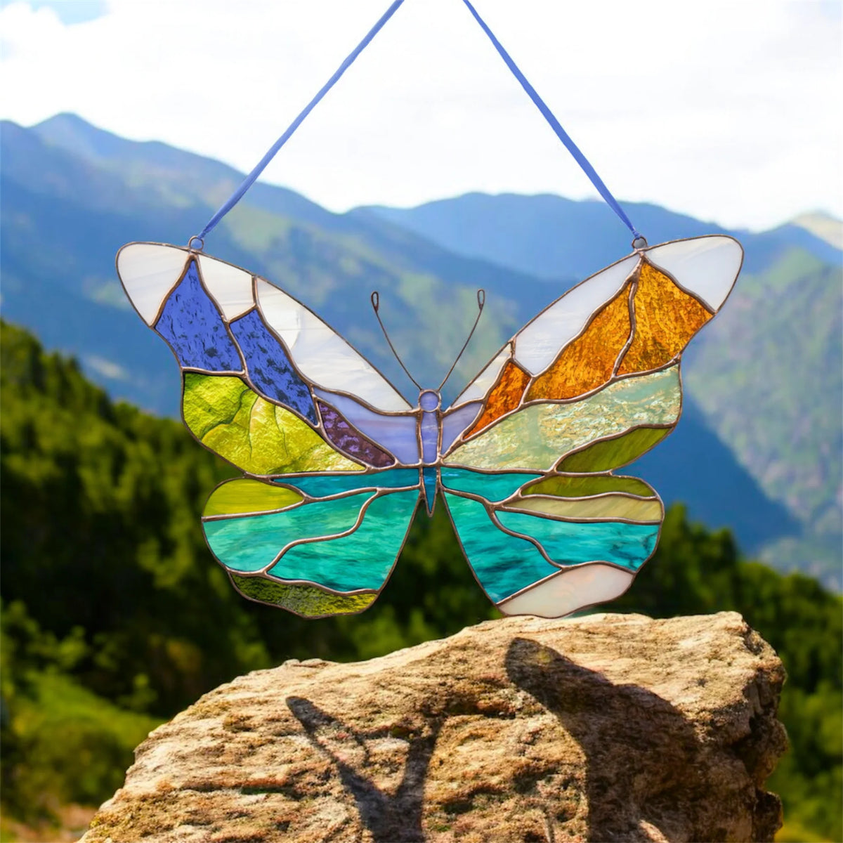 A stained-glass butterfly ornament with multicolored wings hangs from blue strings against a mountain landscape, with a rock in the foreground.