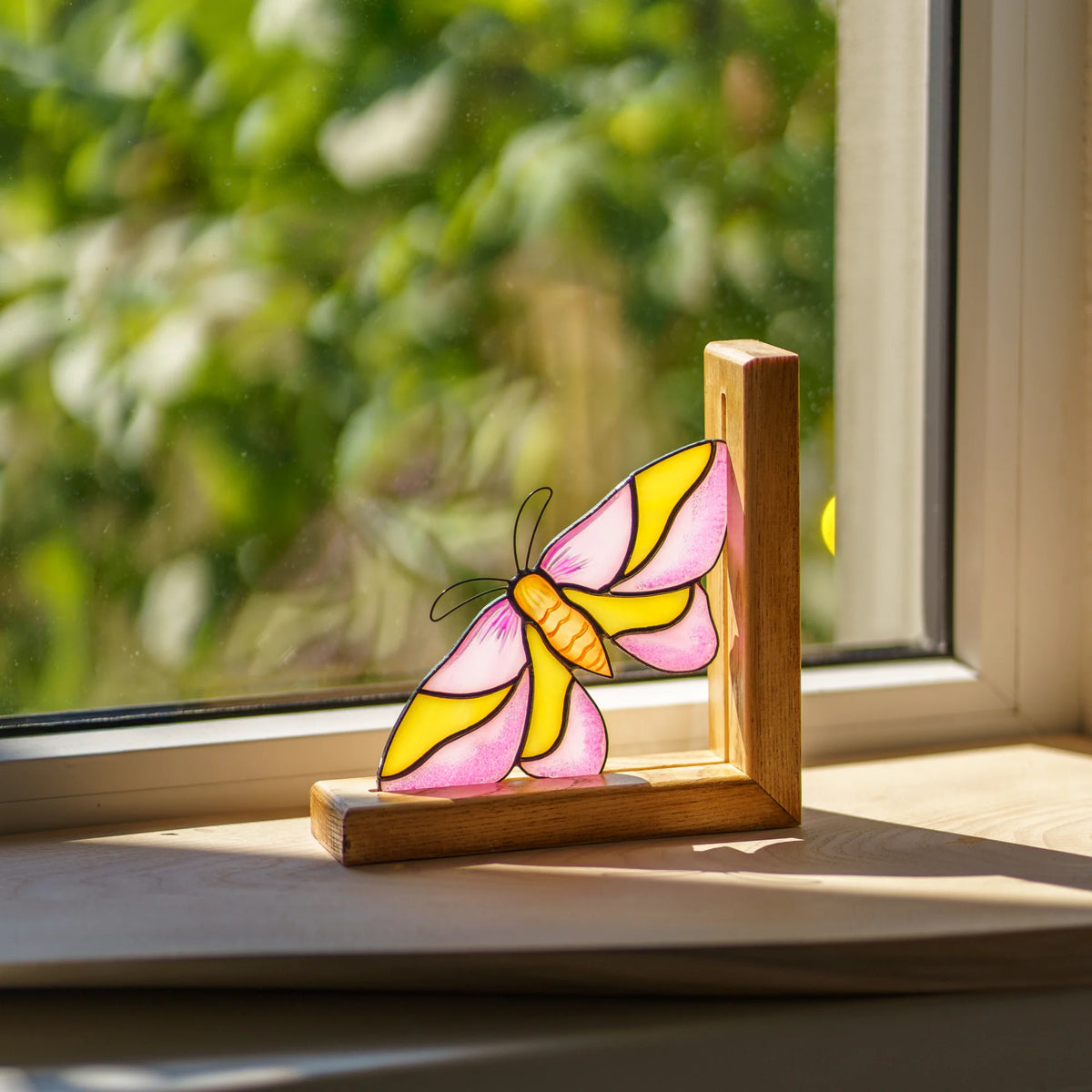 A charming pink and yellow stained glass moth bookend placed on a sunny windowsill, making a wonderful gift for book lovers.