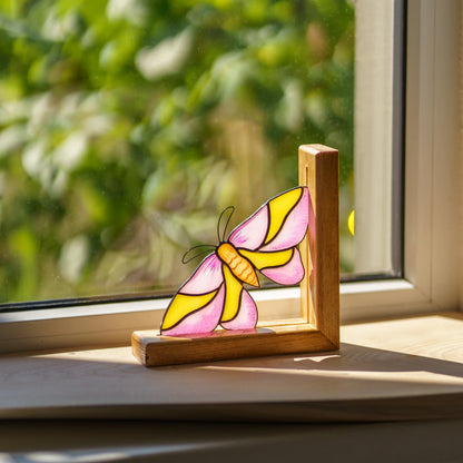A charming pink and yellow stained glass moth bookend placed on a sunny windowsill, making a wonderful gift for book lovers.