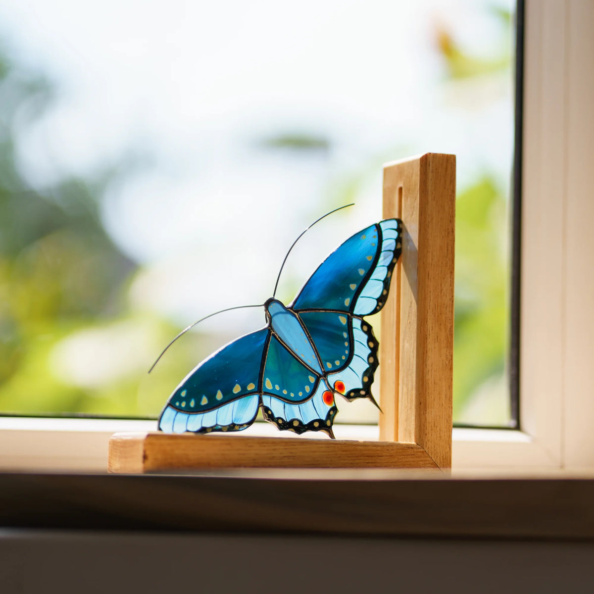 A closer view of the blue stained glass butterfly bookend, highlighting its delicate details and vibrant colors against the natural light of the window.