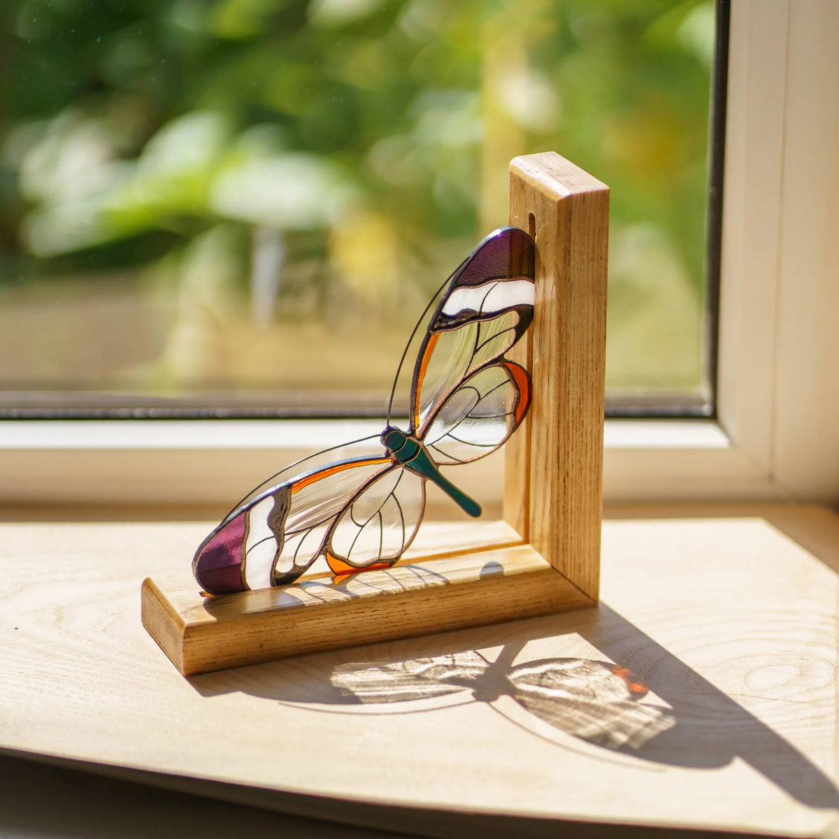 A stained-glass glasswing ornament displayed on a wooden bookend stand on a windowsill, with natural light and a blurred green outdoor background.