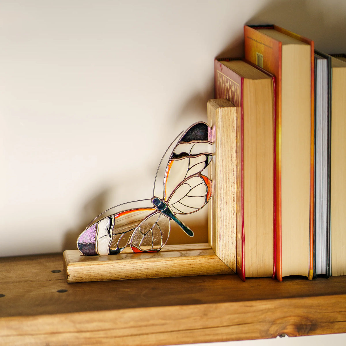 A stained glass glasswing butterfly bookend made of clear glass and metal wire, decorated with colorful glass sections in purple, orange, black, and white, resting on a wooden shelf next to a stack of books.
