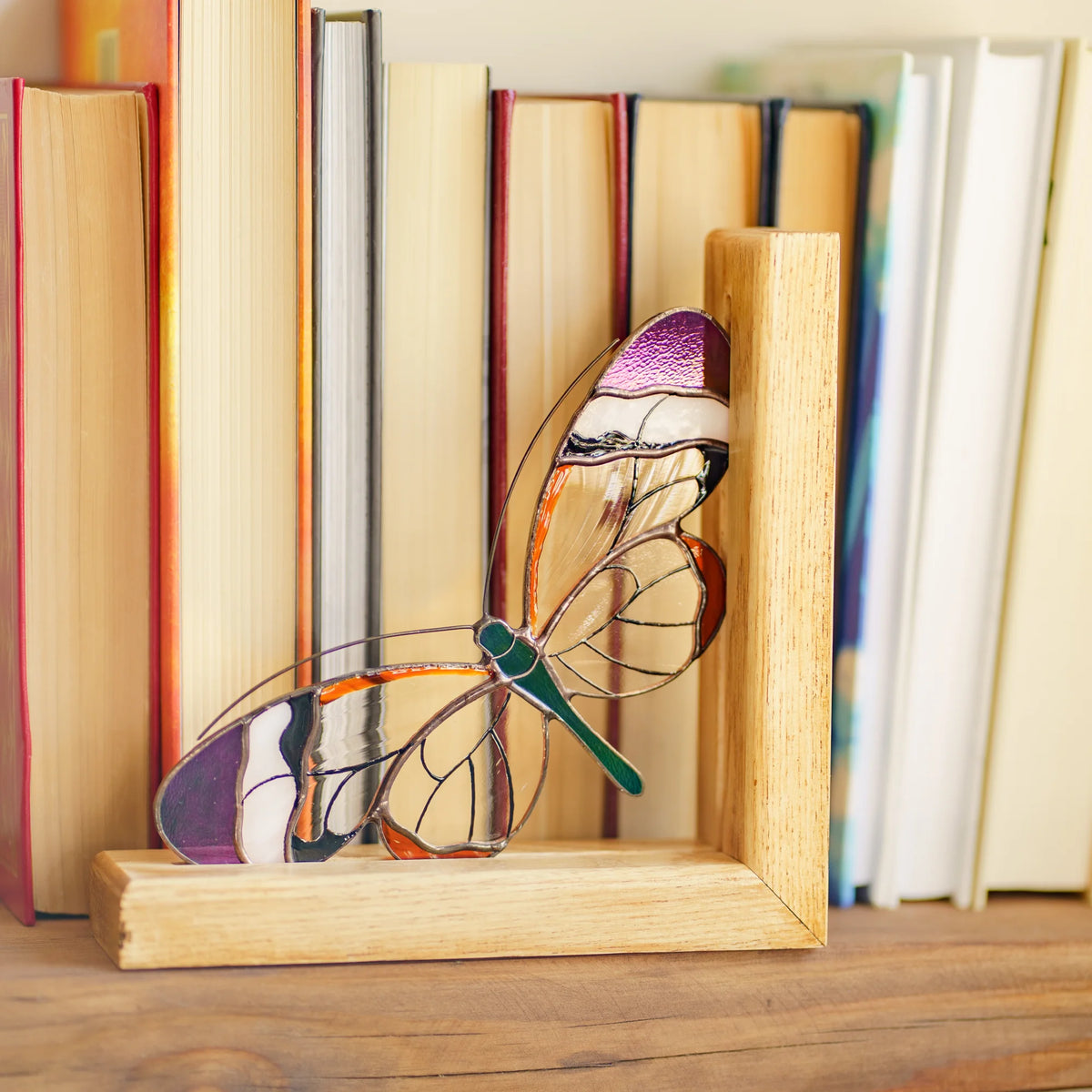 A stained glass glasswing bookend made of light natural wood with a colorful dragonfly design, holding a row of books on a wooden table.