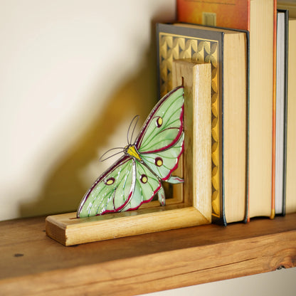 A decorative green moth bookend made from a wooden frame and a stained-glass moth panel, shown on a wooden shelf next to books.