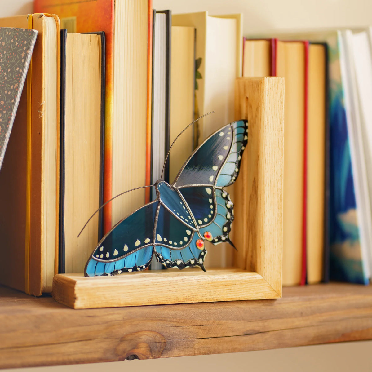 A blue stained glass butterfly bookend placed on a wooden shelf, providing both functional support and a pop of color to your book collection.