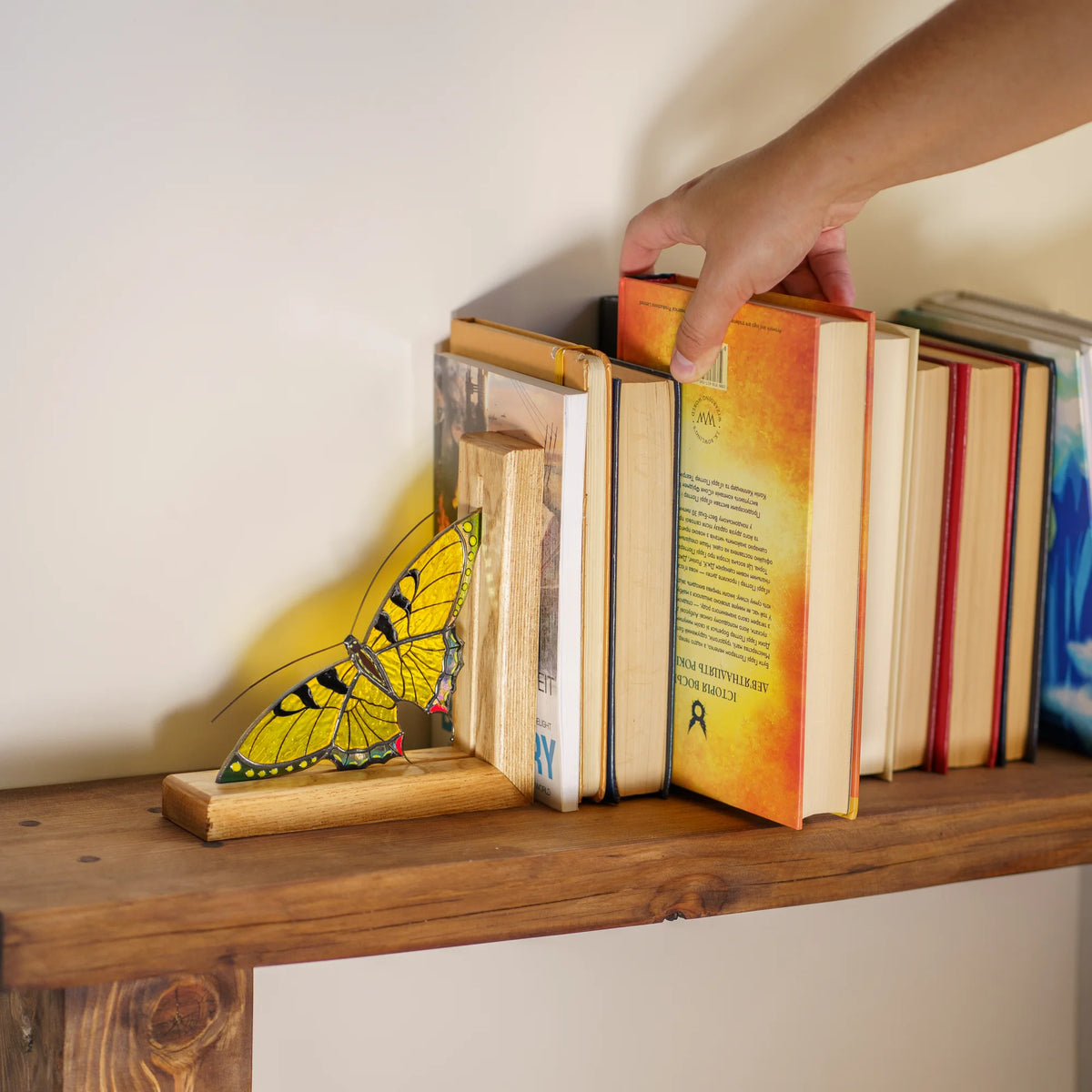 A person placing a book next to a yellow stained glass butterfly bookend on a wooden shelf, showcasing it as a wonderful gift.