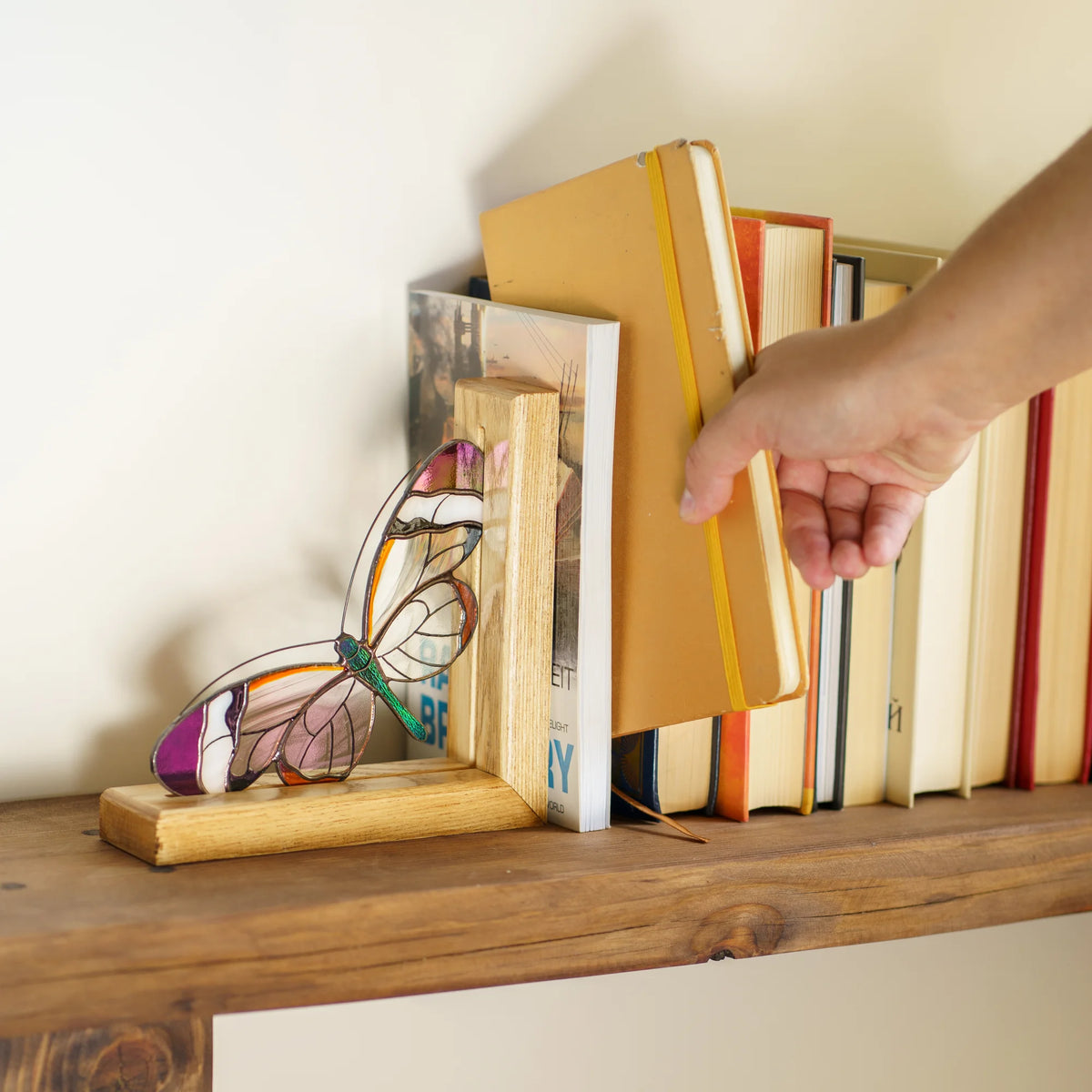 A wooden bookend made with stained glass glasswing butterfly with stained-glass-style wings holds books on a wooden shelf, with a person's hand adjusting one of the books.