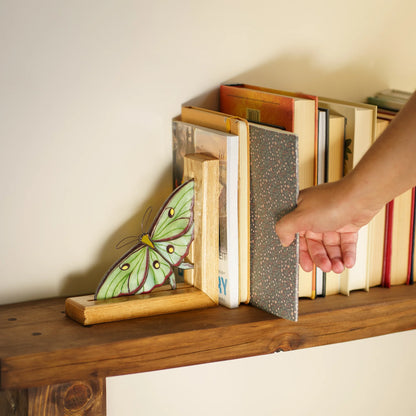 A wooden bookend shaped like a green and yellow luna moth holding a stack of books on a wooden shelf, with a hand adjusting one of the books.