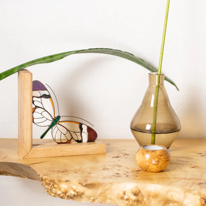 A stained glass butterfly ornament displayed on a wooden stand on a rustic wood slab, next to a glass dropper vase with a green stem against a white background.