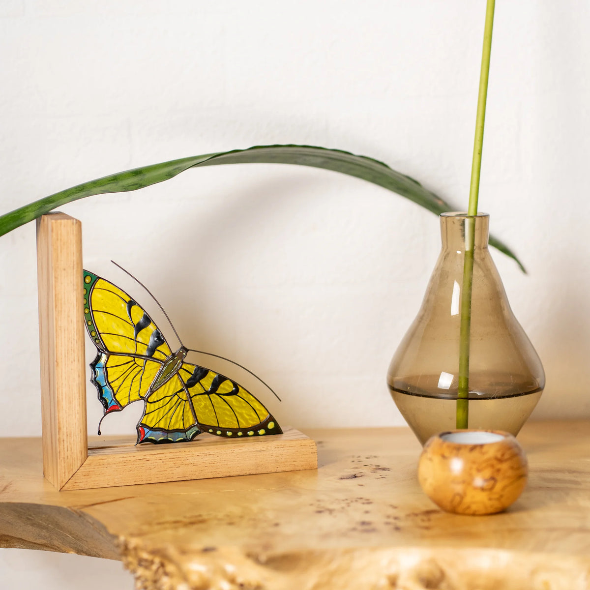 A yellow stained glass butterfly bookend styled on a wooden shelf next to a vase with a plant stem, serving as an exquisite present.