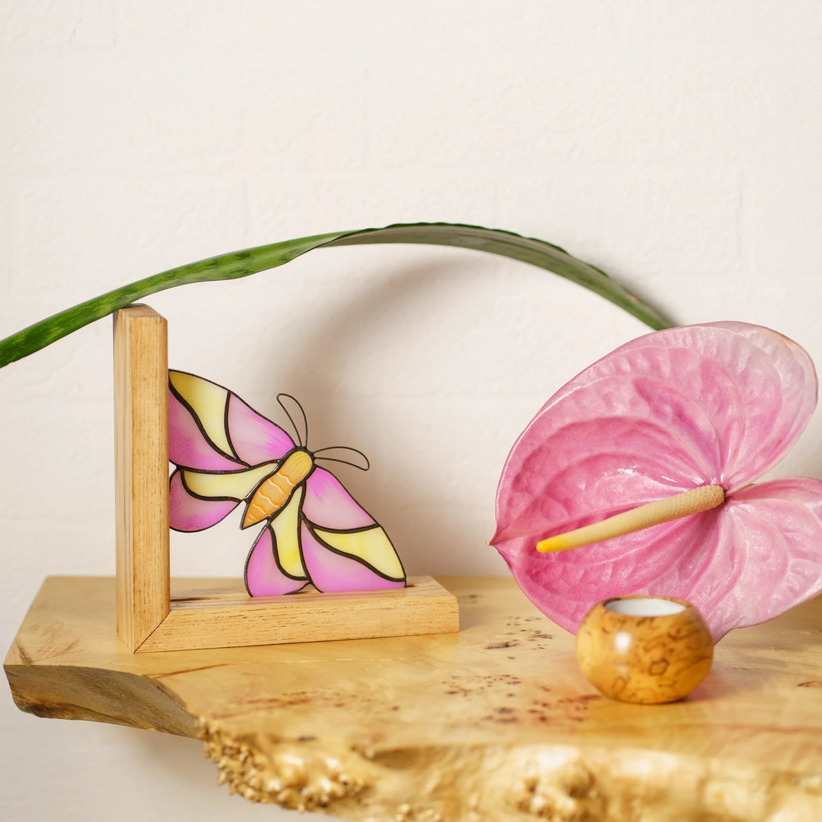 A pink and yellow stained glass moth bookend styled on a wooden shelf next to a pink anthurium flower, serving as an exquisite present.