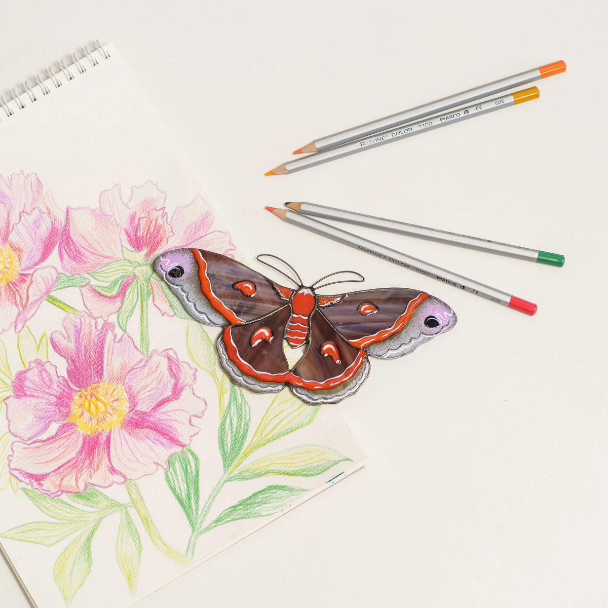 Stained glass cecropia moth suncatcher on a sketchbook with peony drawings and colored pencils