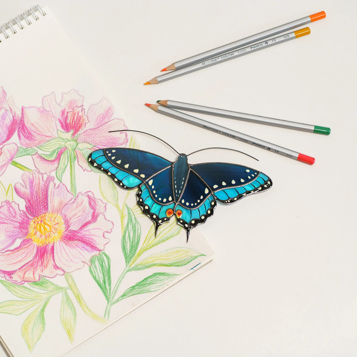 Blue swallowtail butterfly stained glass suncatcher with orange eyespots, beside a peony sketch and colored pencils