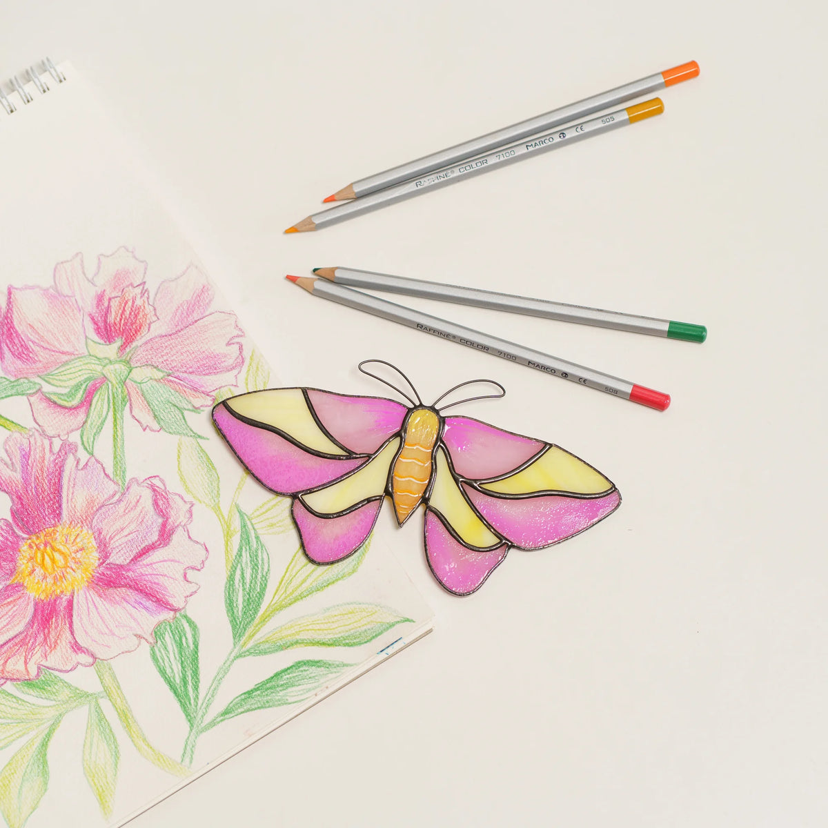 Rosy maple moth suncatcher in pink and yellow stained glass, laid on a peony sketch next to colored pencils