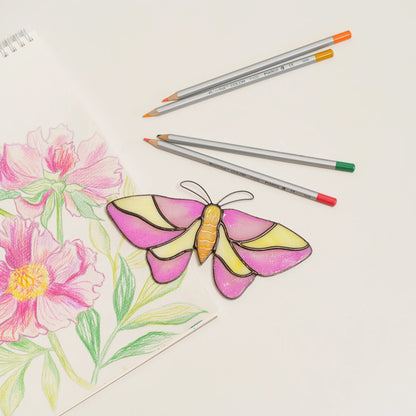 Rosy maple moth suncatcher in pink and yellow stained glass, laid on a peony sketch next to colored pencils