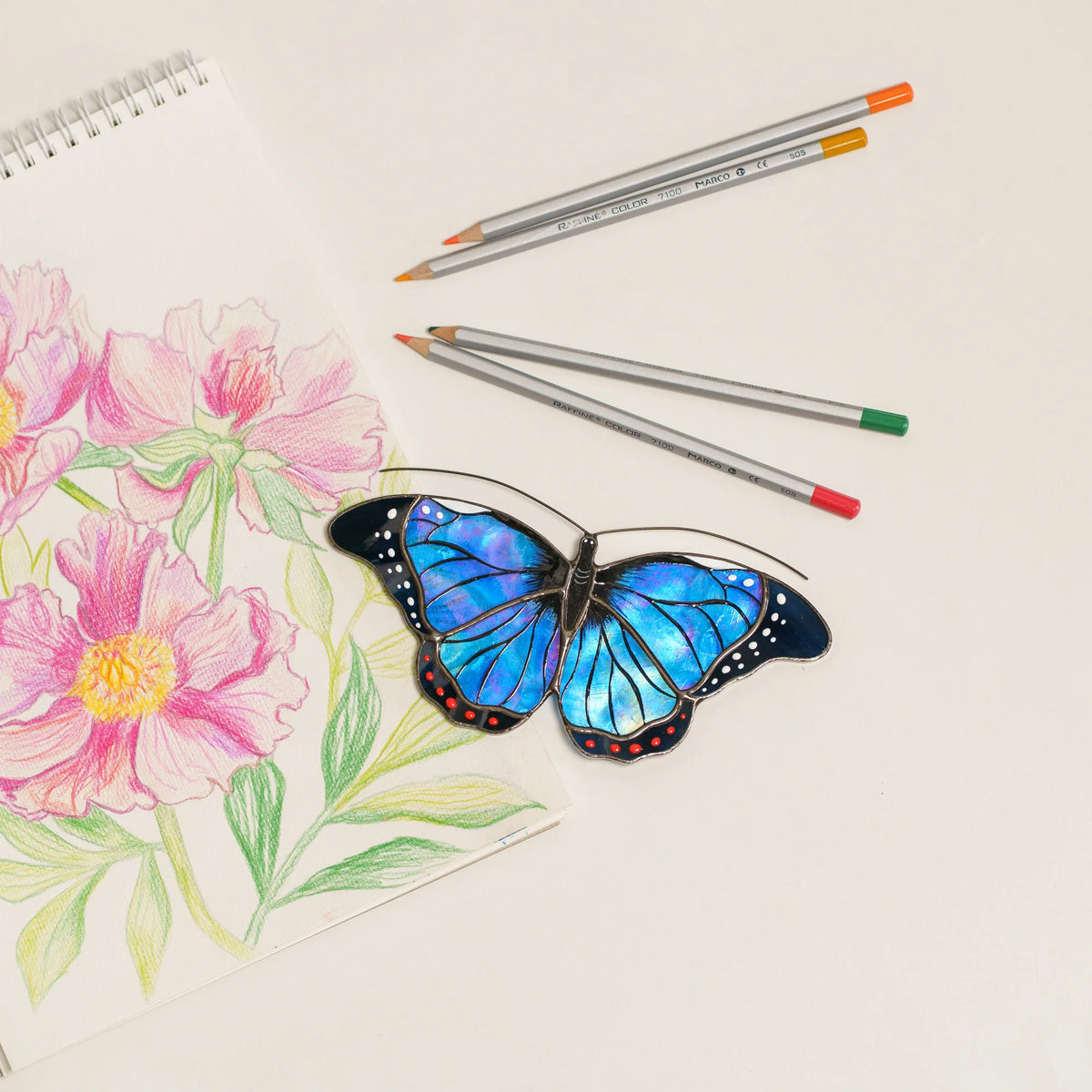 Blue morpho stained glass suncatcher with iridescent blue wings and red dots, beside colored pencils