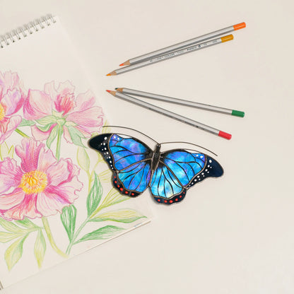 Blue morpho stained glass suncatcher with iridescent blue wings and red dots, beside colored pencils