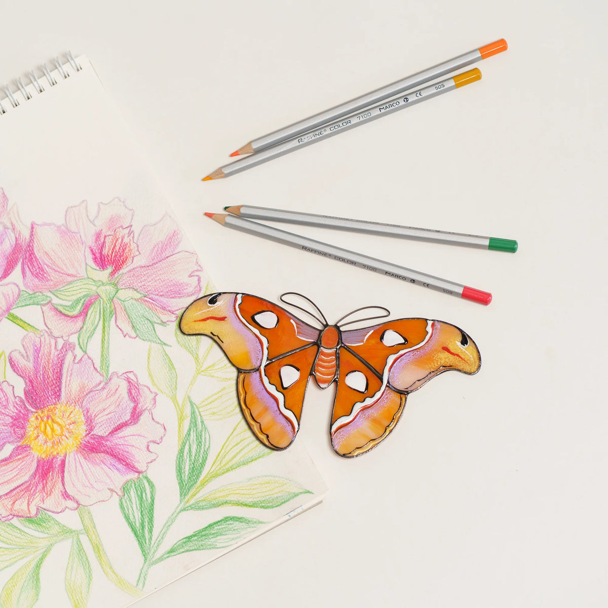 Atlas moth stained glass suncatcher in orange and lilac glass with snake-head wingtips, on a peony sketch