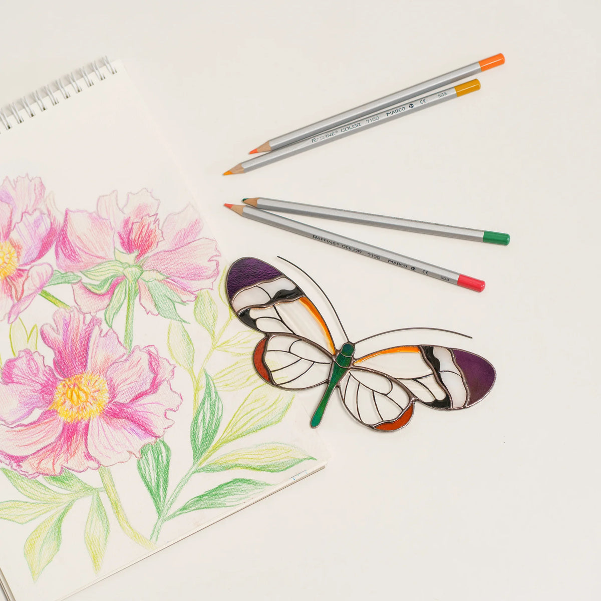 Glasswing butterfly suncatcher with clear wings, purple tips and a green body, on a sketchbook with pencils