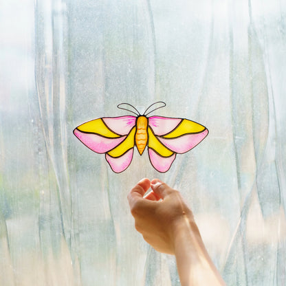 Rosy maple moth suncatcher on a bright window, pink and yellow glass glowing above a reaching hand