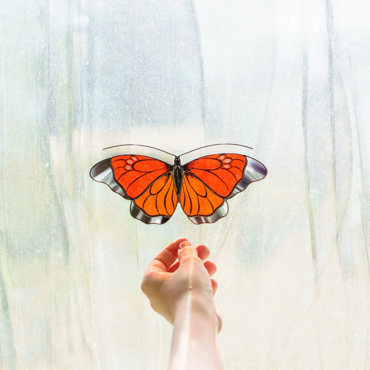 Orange butterfly suncatcher glowing in a window as a hand reaches toward it
