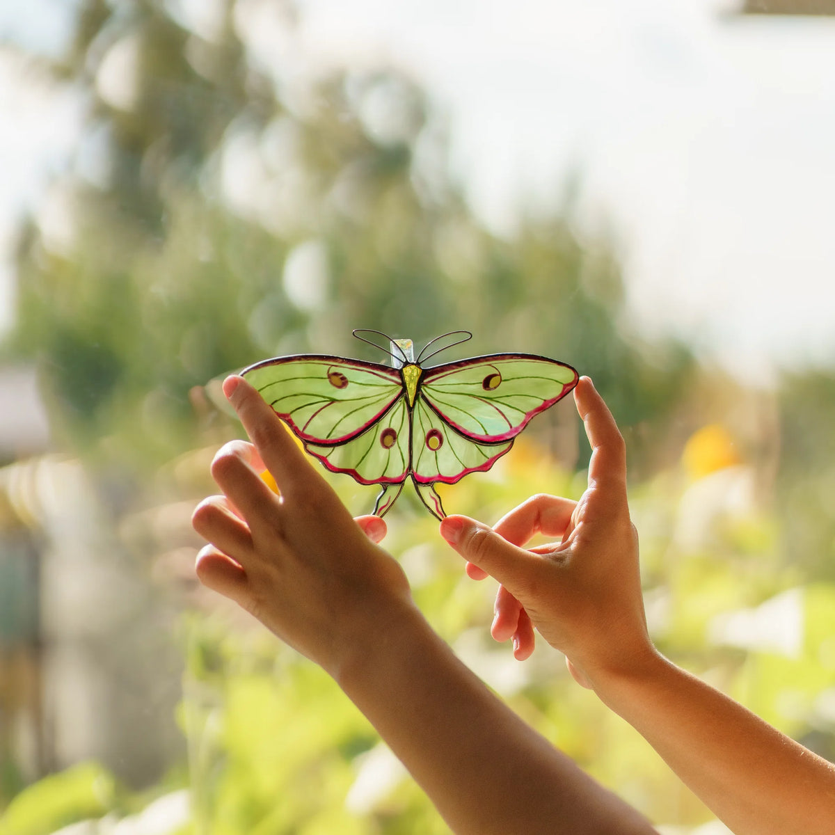 Two hands framing a luna moth stained glass suncatcher against a sunlit garden window