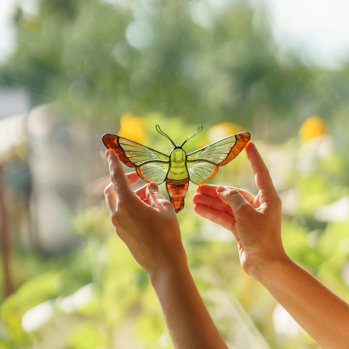 Hands holding a hummingbird moth suncatcher in green and amber glass against a blurred summer garden