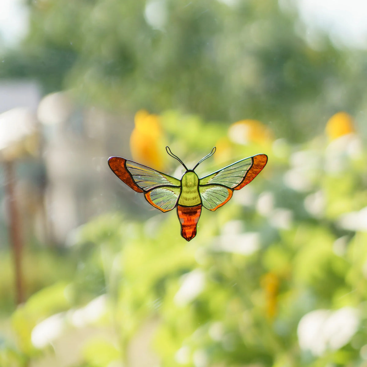 Hummingbird moth stained glass suncatcher on a window, amber wingtips lit by sun from the garden outside