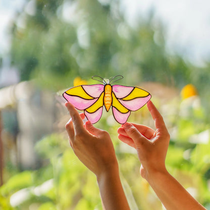 Hands holding a rosy maple moth suncatcher in pink and yellow glass up to bright garden light