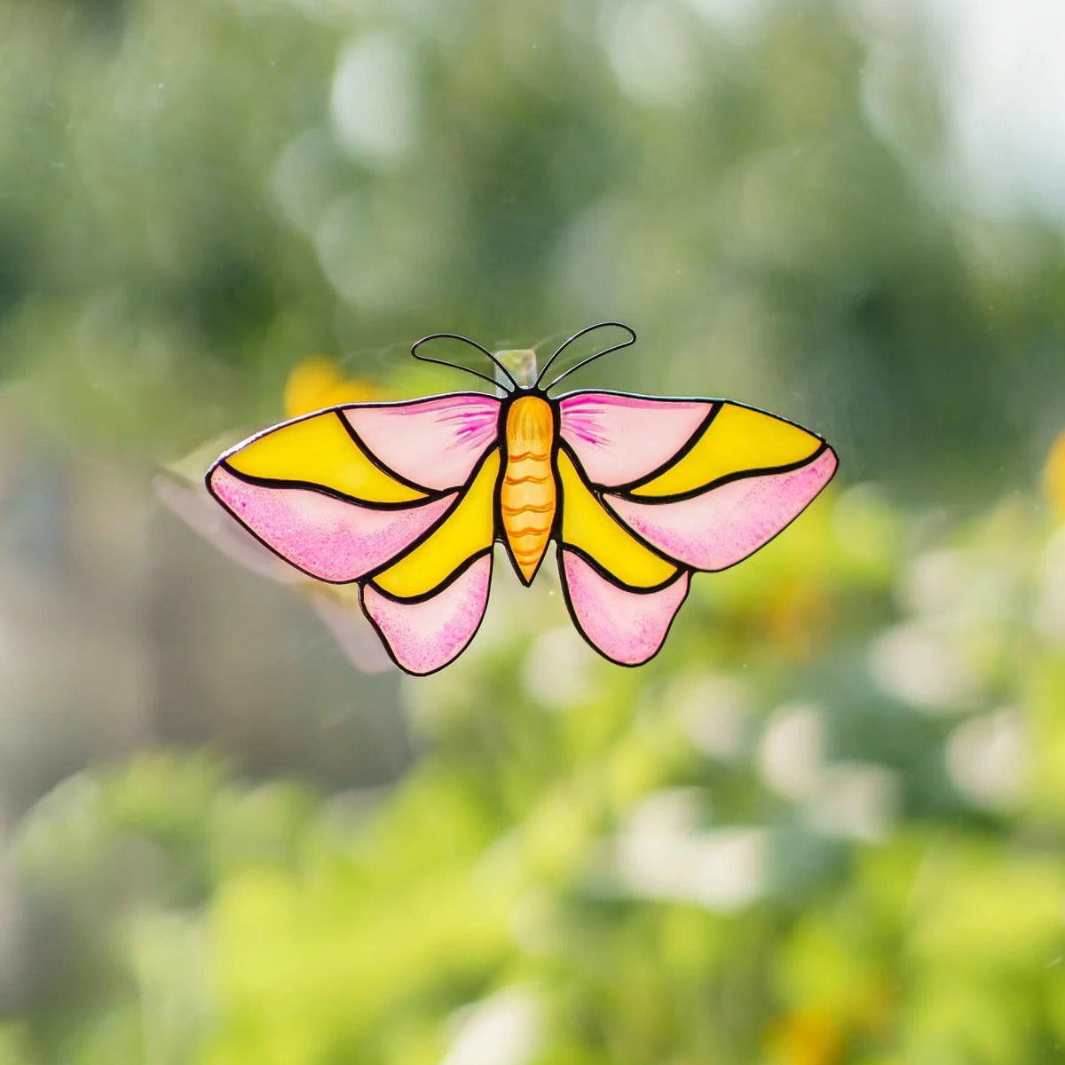Rosy maple moth stained glass suncatcher on a window above a blooming summer garden