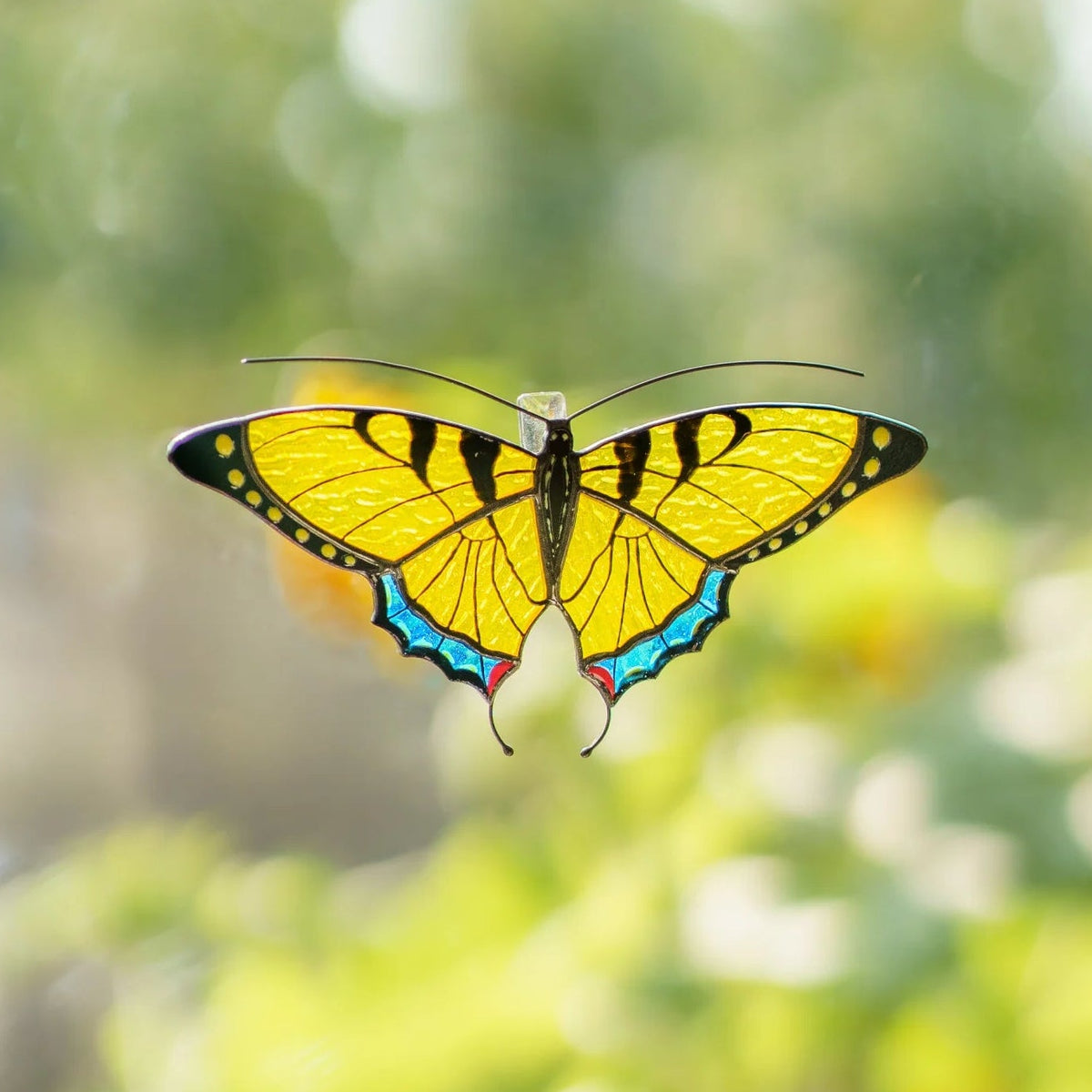 Yellow swallowtail stained glass suncatcher on a window with a sunflower garden blurred behind it