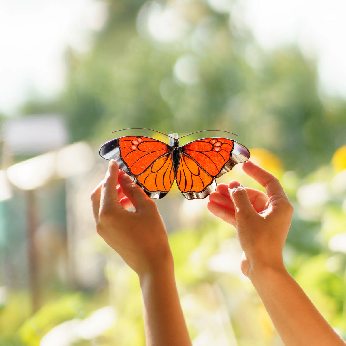 Two hands holding an orange butterfly suncatcher toward the sun, red-orange glass bright against the garden