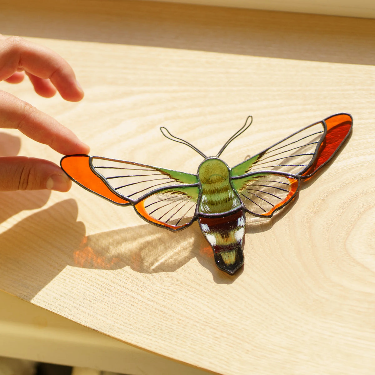 Hummingbird moth stained glass suncatcher on a sunny wooden windowsill, casting amber light across the wood